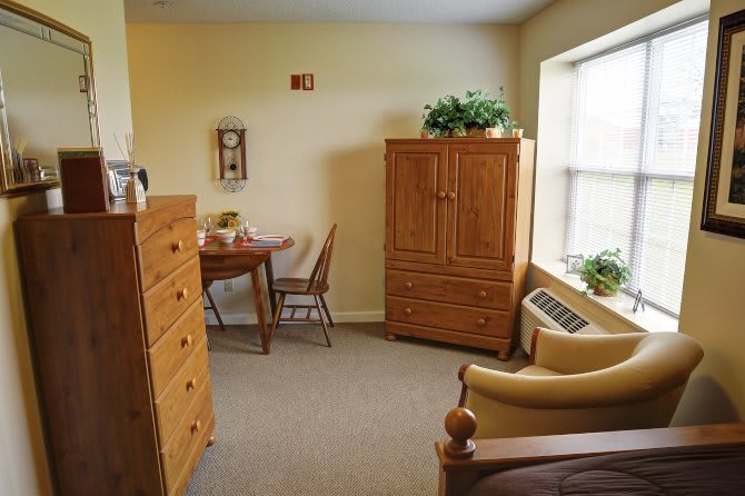 A cozy furnished room with a wardrobe, dresser, table and chairs, indoor plants, and an armchair by the window.