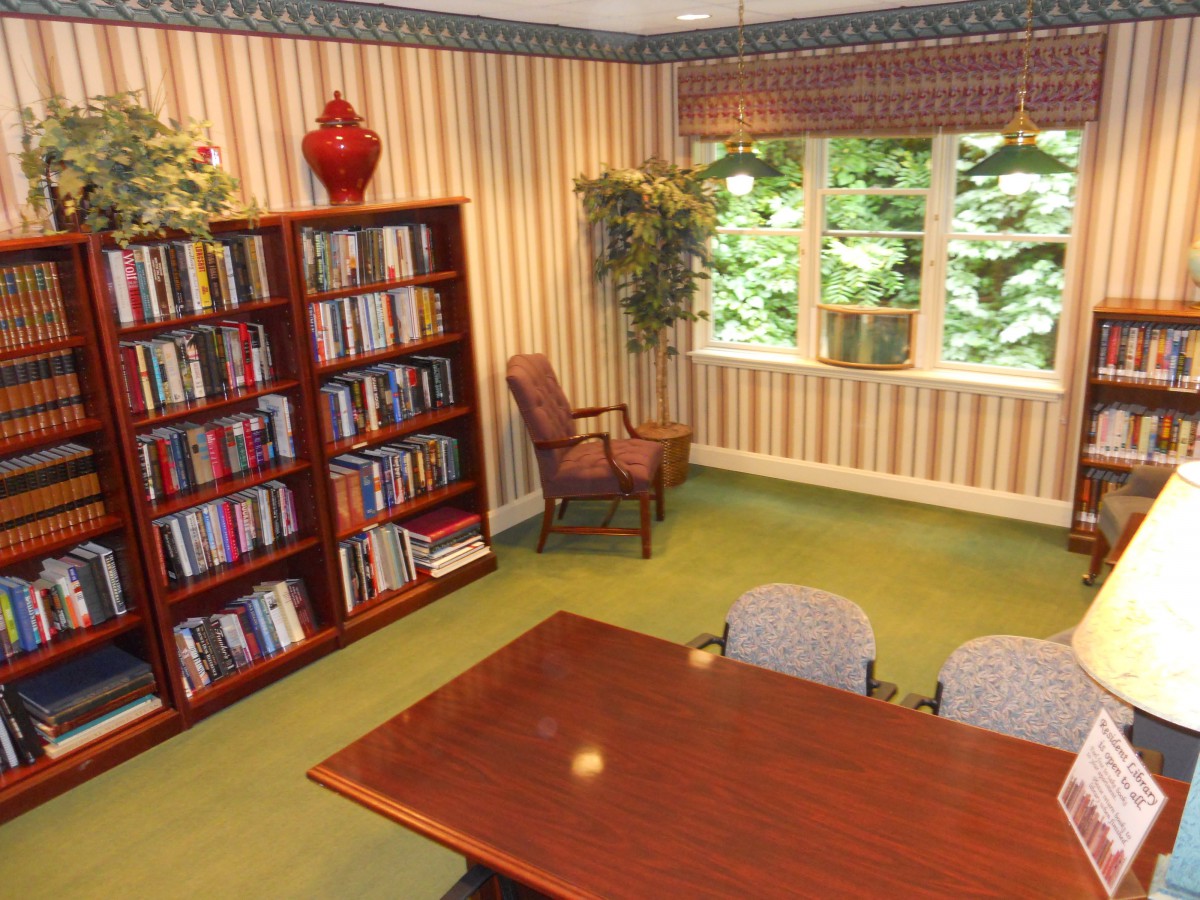 A cozy library space with a bookshelf, table, and chairs near a window with a view of greenery.