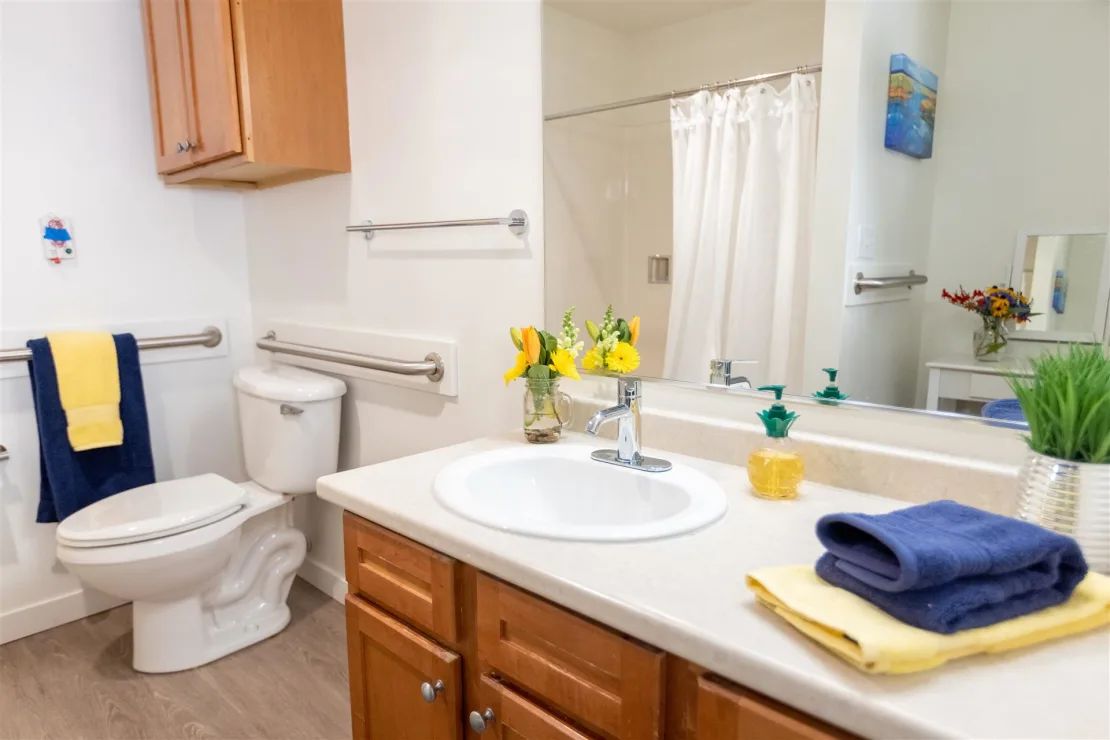 A clean and neatly organized bathroom with a sink, mirror, toilet, and a shower adorned with colorful towels and flowers.