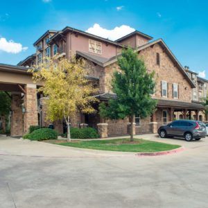 A residential building with a brick facade, surrounded by trees and a car parked on the driveway under a clear blue sky.
