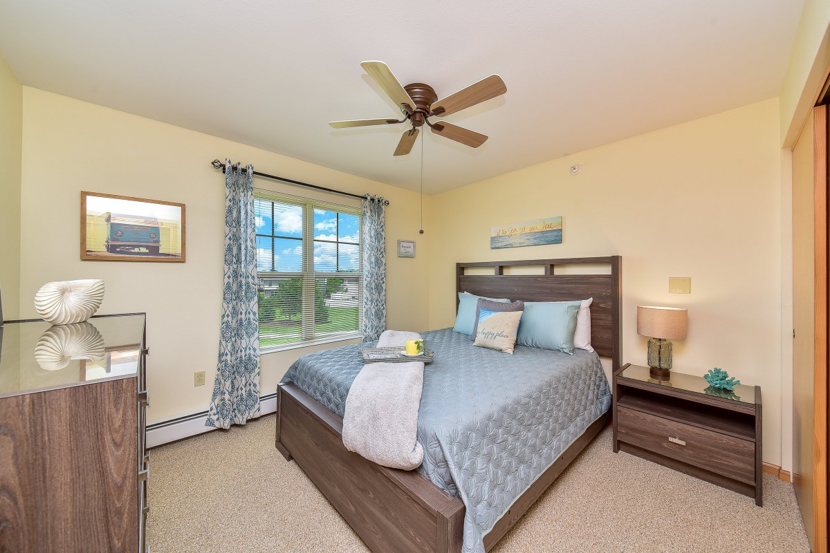 A cozy bedroom with a neatly made bed, ceiling fan, and large window with patterned curtains.