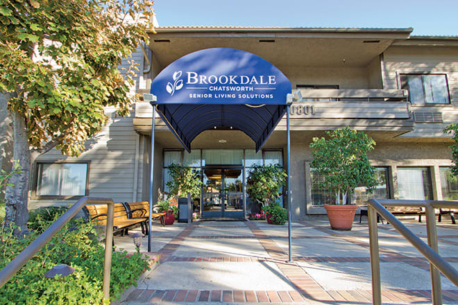 Entrance to a building with a blue canopy marked 'Brookdale', surrounded by plants and benches.
