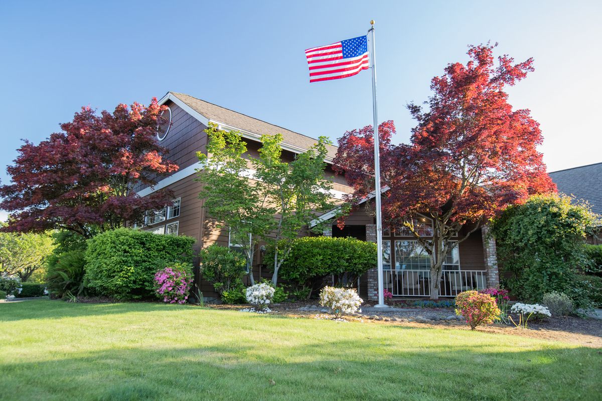A suburban house with an American flag flying in the yard, surrounded by a well-maintained garden and trees.