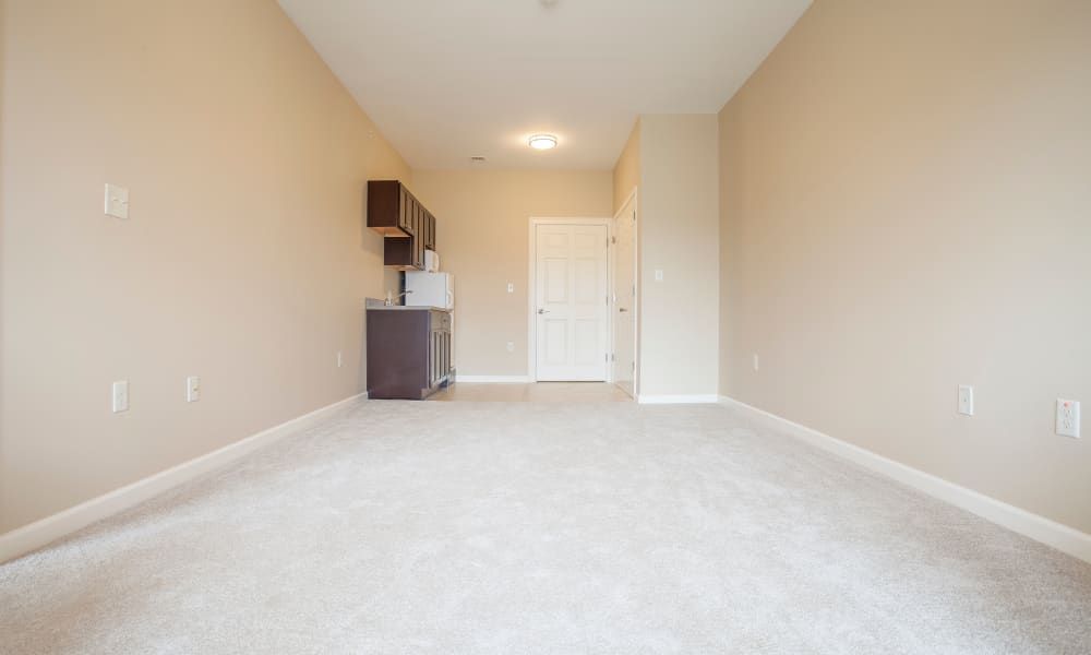 A minimalist, unfurnished apartment with a kitchenette and carpeted flooring.