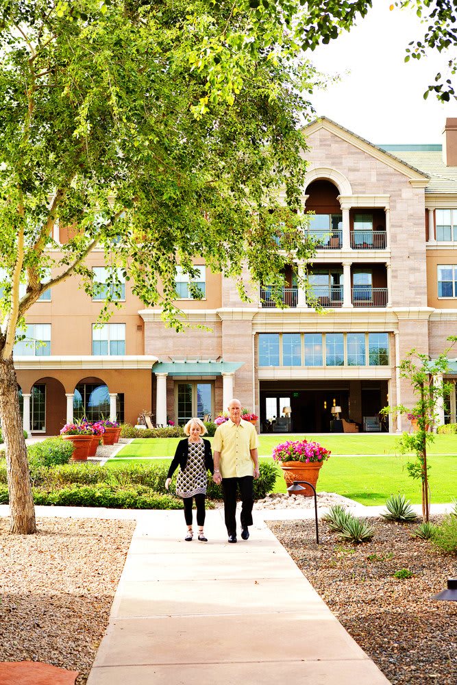 An elderly couple walking towards a retirement home surrounded by a garden.