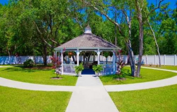 A charming white gazebo is centered in a neatly landscaped garden, surrounded by lush green grass and trees, with pathways leading up to it.
