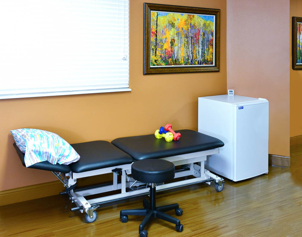 A therapy room featuring a treatment table with a pillow, a stool, a colorful toy, and a painting on the wall. There is also a small fridge in the corner of the room.