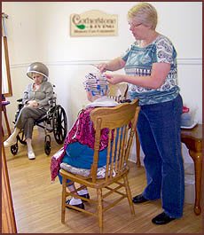 An elderly woman sits in a wheelchair while another receives a haircut in a community care setting.