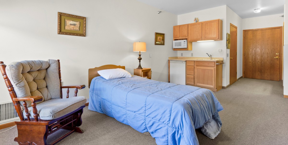 A cozy bedroom with a single bed, blue bedding, and a kitchenette in the corner.