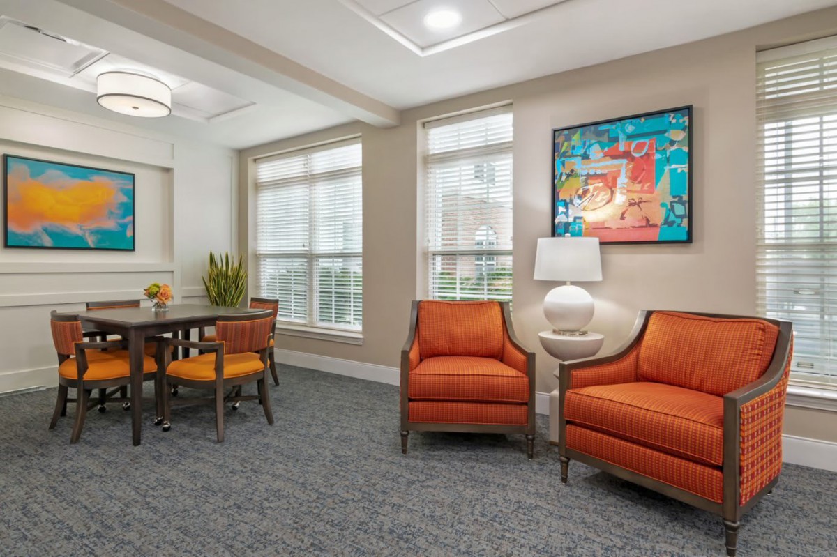 A cozy living room setting featuring orange chairs, a table with seating, artwork on the wall, and large windows that allow ample natural light.