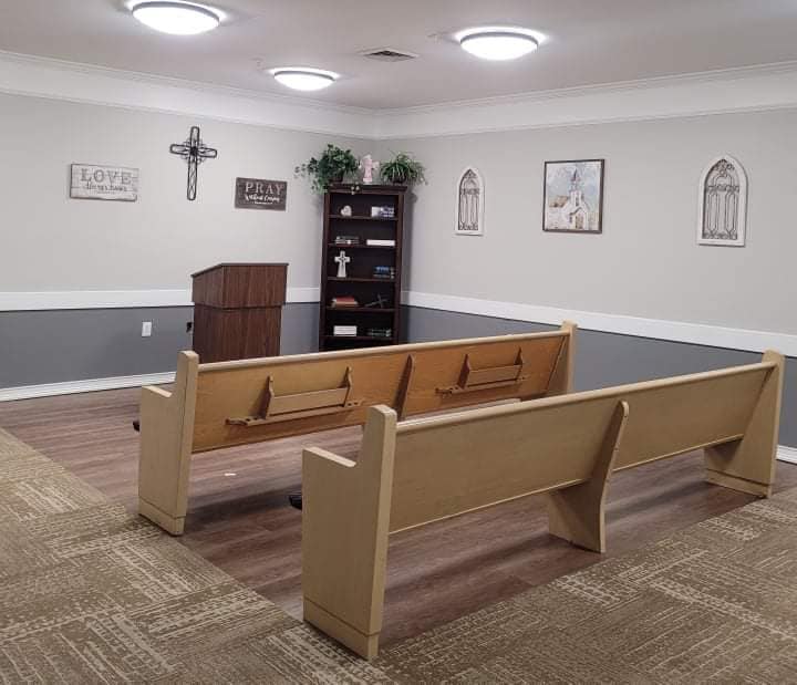 A small chapel with wooden pews, a cross on the wall, and bookshelves.