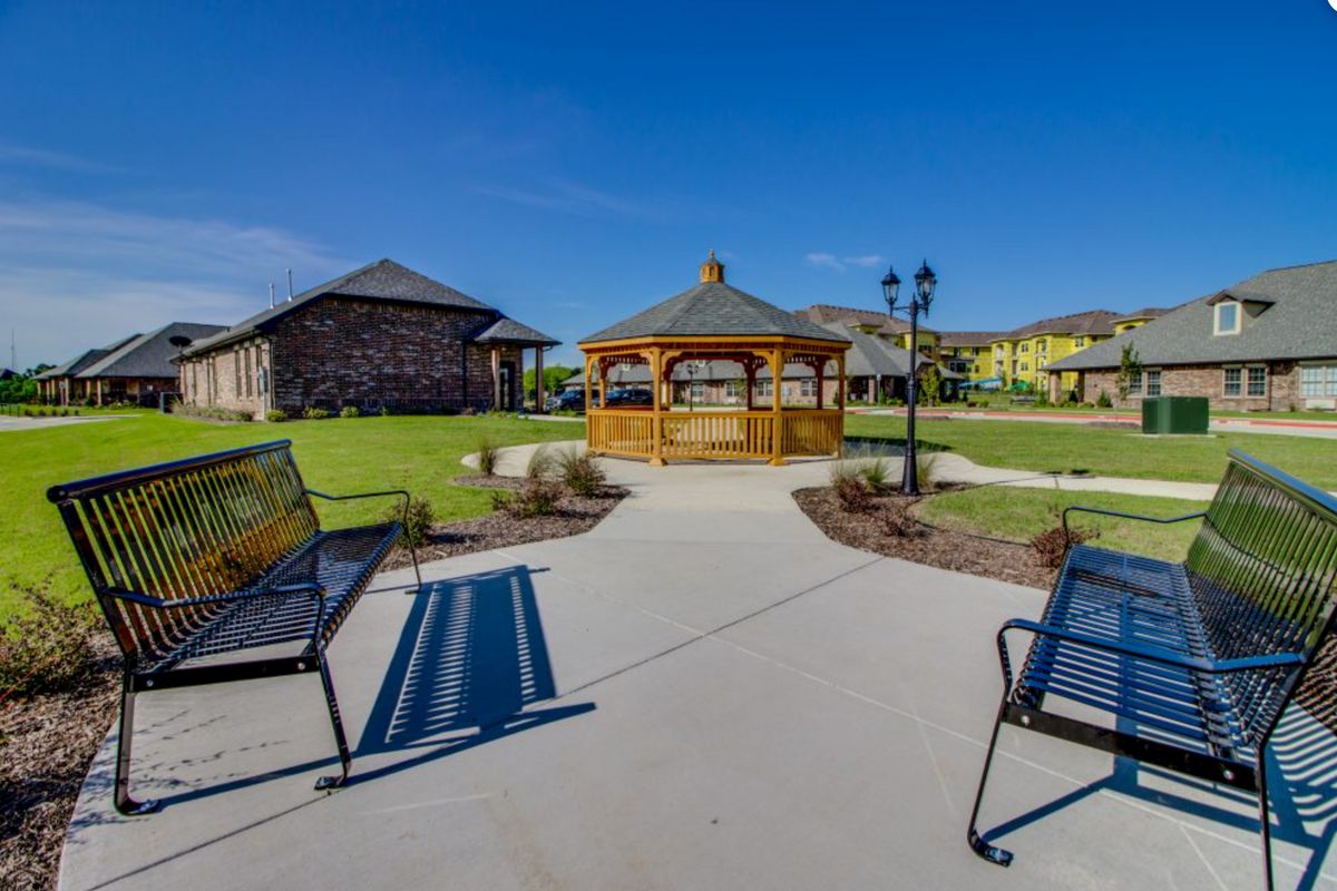 A picturesque outdoor area featuring a gazebo surrounded by benches, a well-maintained lawn, and adjacent buildings under a clear blue sky.