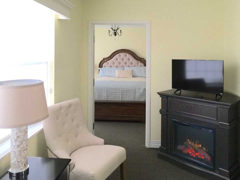 A cozy bedroom with a TV and fireplace, featuring a plush chair and a bed with an ornate headboard.