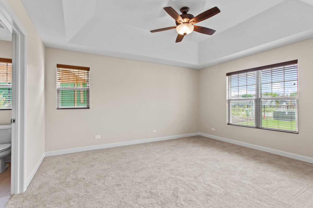 An empty room with a ceiling fan and carpeted floor, featuring large windows that allow natural light.
