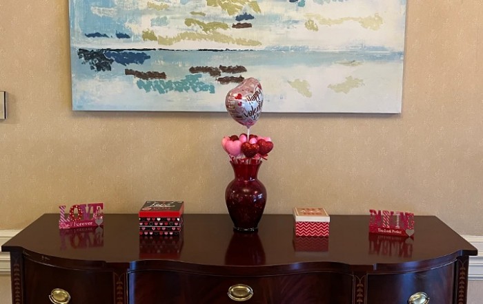 A decorative arrangement with a red vase, flowers, and a balloon on a wooden table beneath a painting.