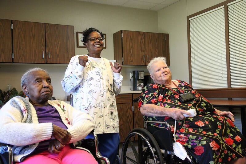 A caretaker stands with two elderly individuals in wheelchairs at a nursing home.