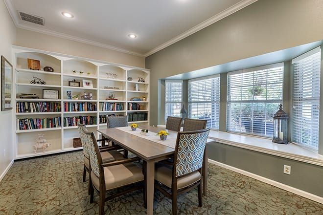 A cozy library with bookshelves, a table surrounded by chairs, and a large window letting in natural light.