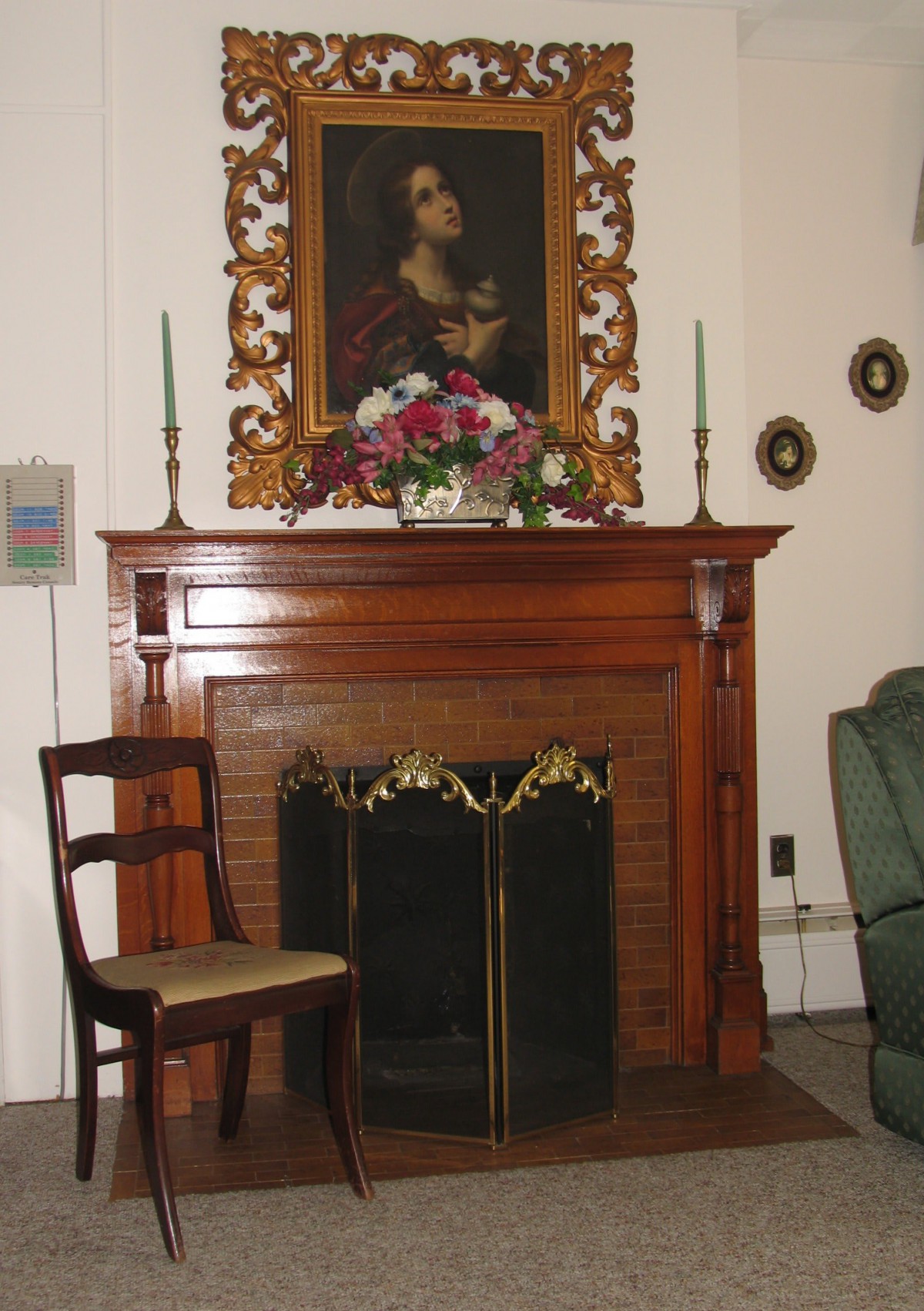 A classic indoor setting featuring a wooden fireplace mantel adorned with candles, a floral arrangement, and a portrait. A single chair rests at the side.
