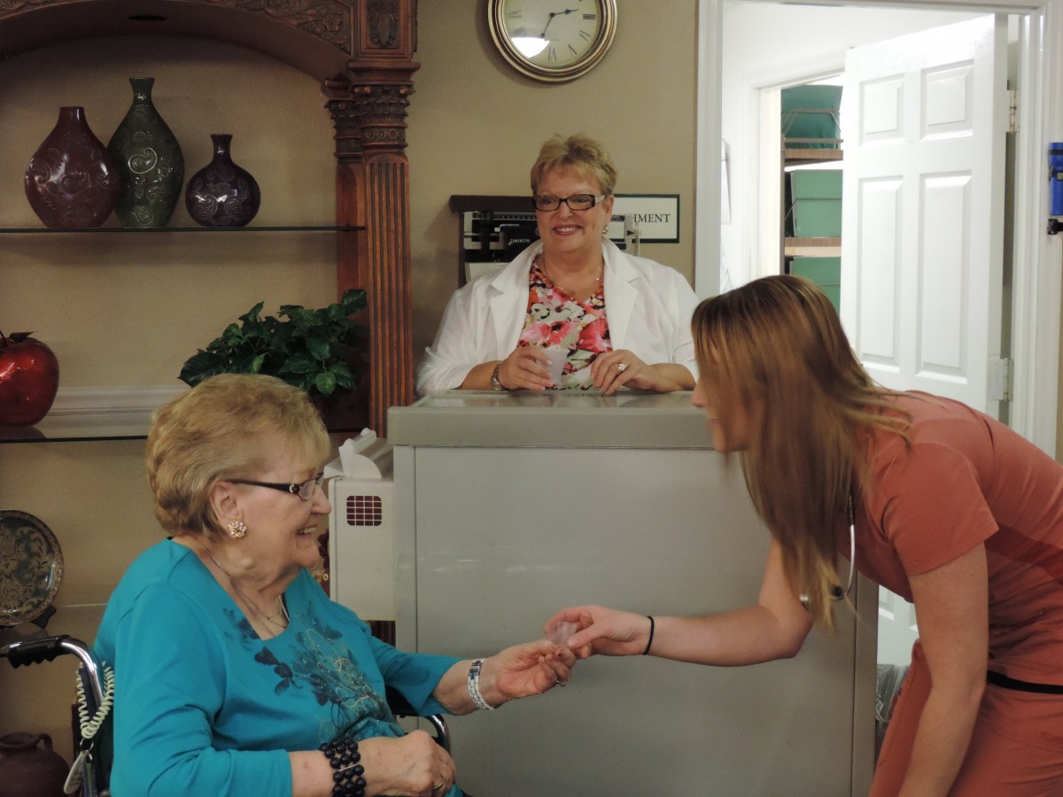 An elderly woman sits in a reception area as another woman leans in to interact with her, both smiling.