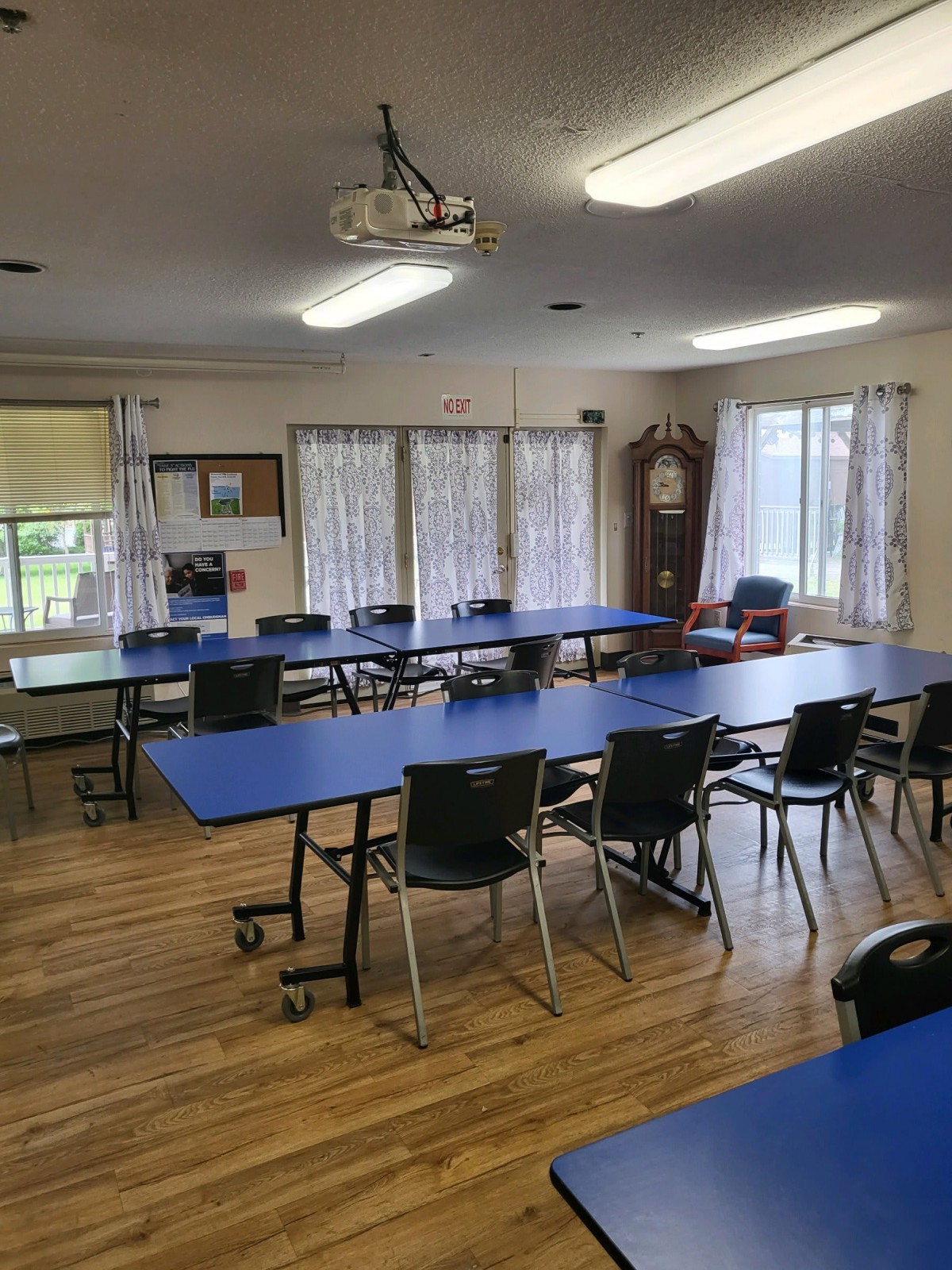 An empty classroom or meeting room with blue tables and chairs, featuring large windows with curtains and a clock in the corner.