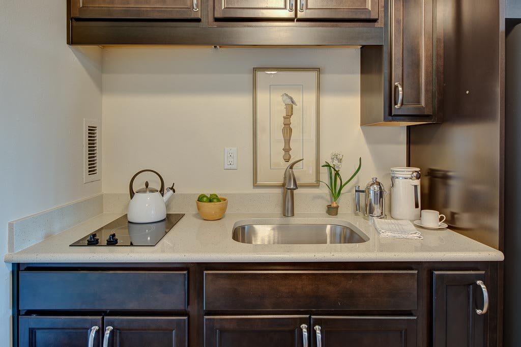 A compact kitchen with dark cabinets, featuring a countertop with a sink, tea kettle, and minimalist decor.