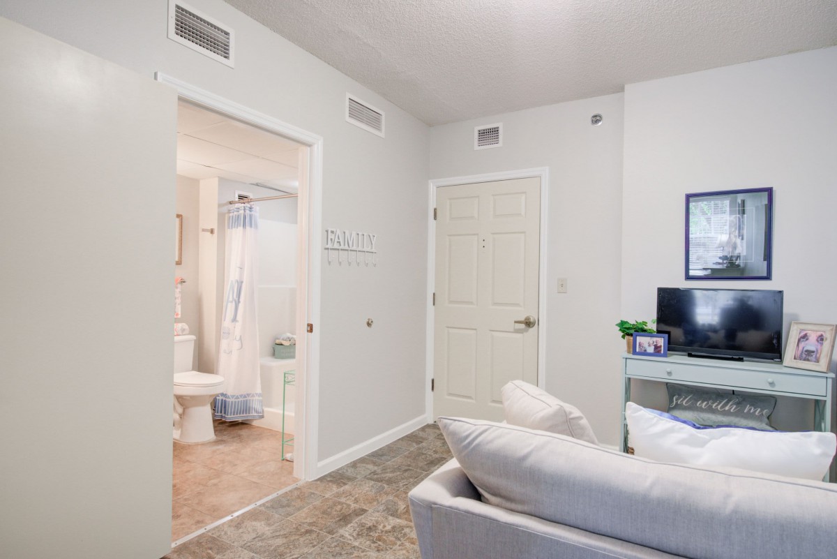 A modern living room with a view into an adjoining bathroom, featuring a comfortable sofa, desk with computer, and neutral color scheme.