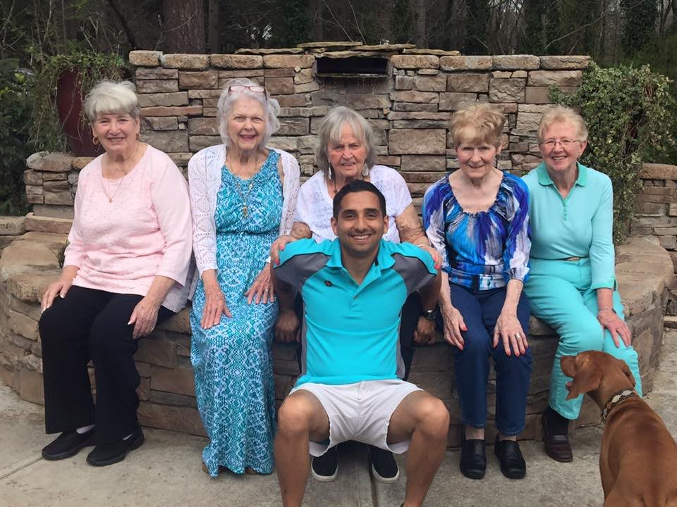 A group of elderly women and a young man sitting and smiling together outdoors.