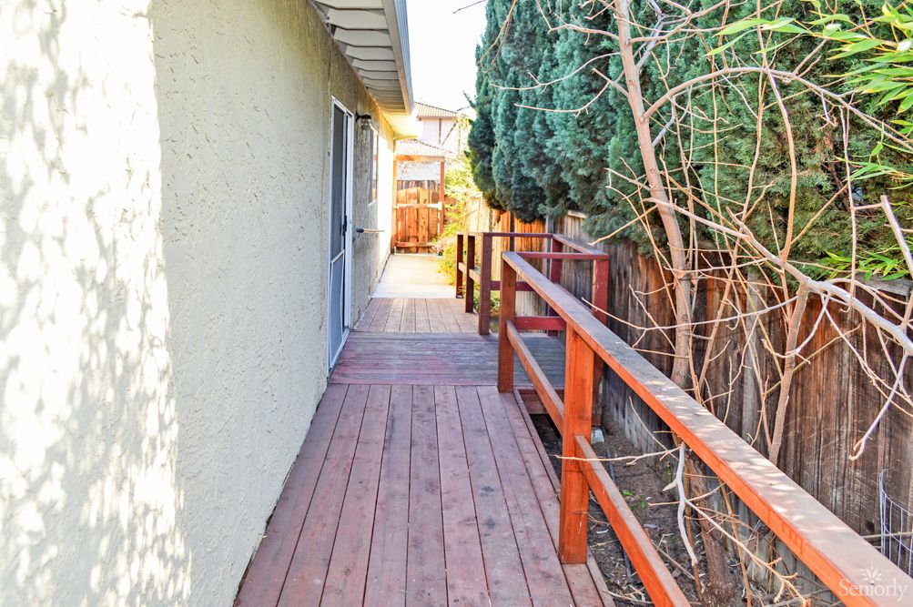 A wooden deck pathway with railings alongside a house, bordered by trees and a fence under sunlight.