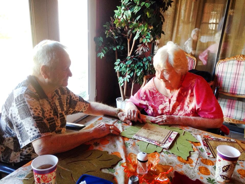An elderly couple holding hands at a table, surrounded by warm lighting and plants.