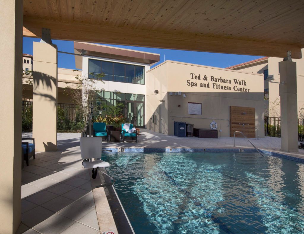 An outdoor swimming pool area adjacent to a fitness and spa center on a sunny day.