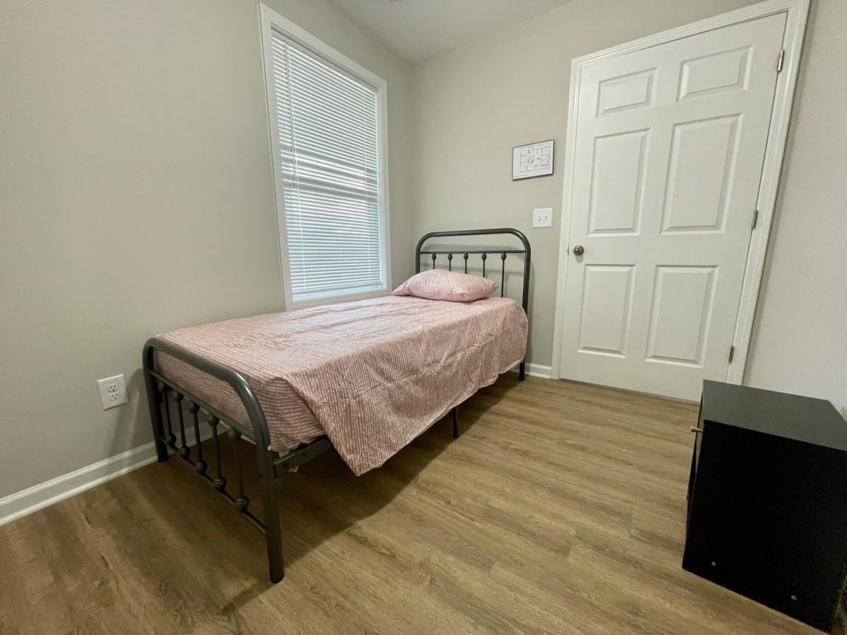 A simple bedroom with a single metal-framed bed and pink bedding, featuring a window and wooden flooring.