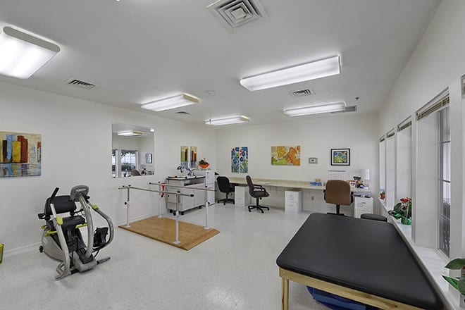 A clean and well-equipped physical therapy room featuring an exercise bike, parallel bars, and a therapist's desk.