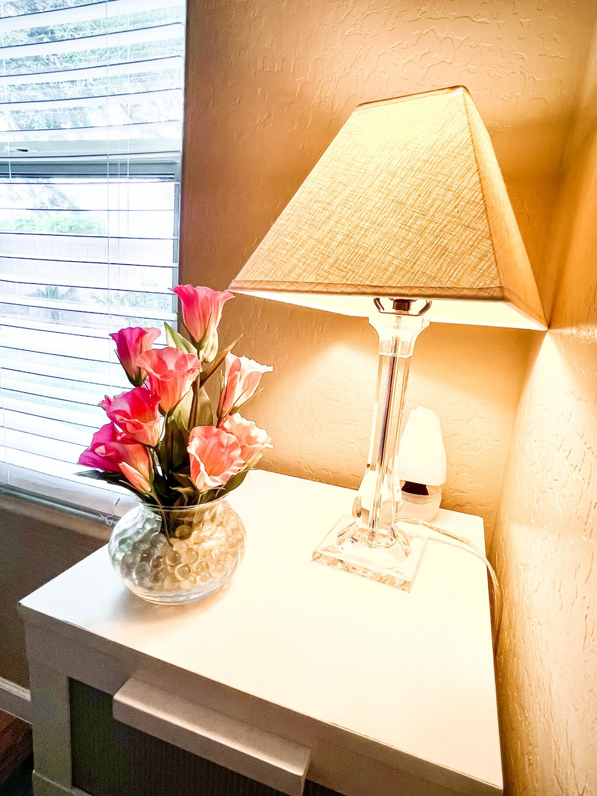 A cozy setup with a lamp and pink flowers in a vase on a table by the window.
