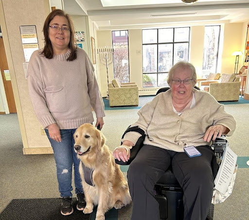 A heartwarming moment shared between a woman in a wheelchair and her companion dog, alongside a caregiver in a cozy indoor setting.