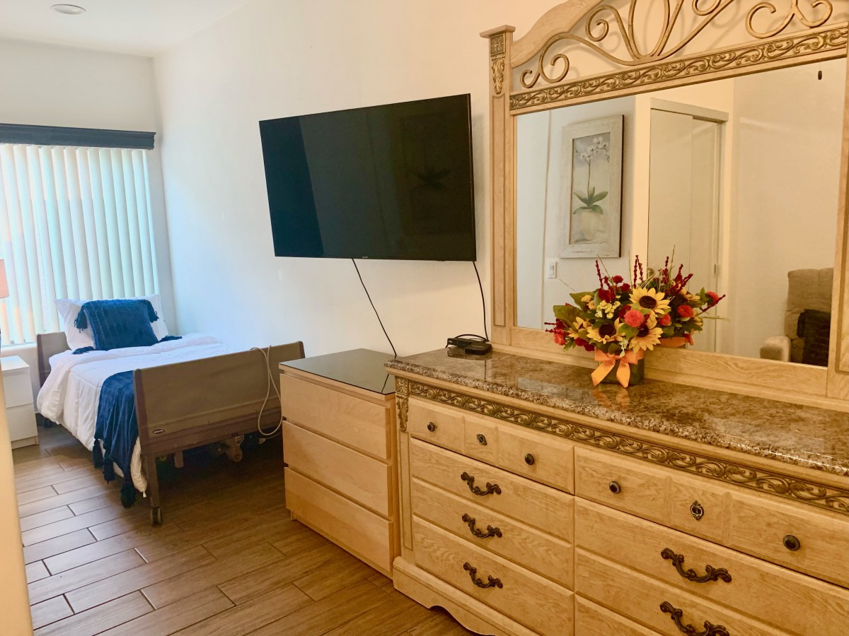 A cozy bedroom featuring a dresser with a mirror, a mounted television, and a vibrant flower arrangement.