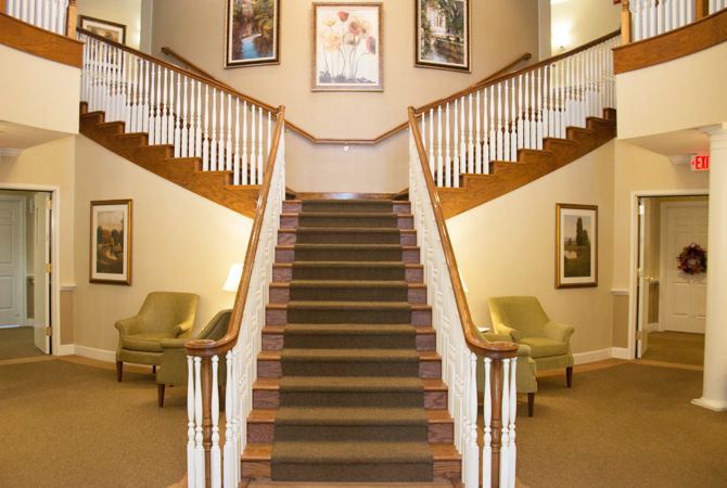 A grand staircase with wooden railings and carpeted steps, flanked by chairs and decorated with artwork.