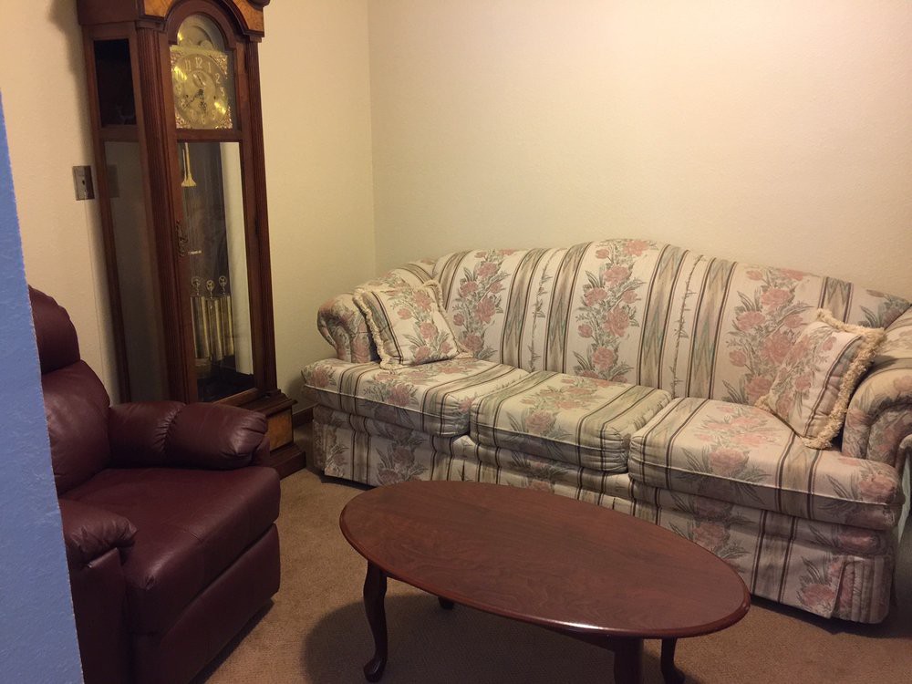 A cozy living room with a patterned sofa, leather armchair, wooden coffee table, and a grandfather clock in the corner.