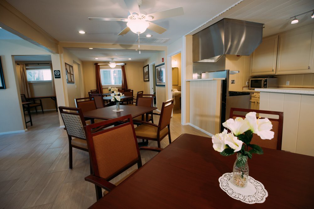 A cozy dining area with wooden tables and chairs, featuring a kitchen in the background and decorative flowers on the table.