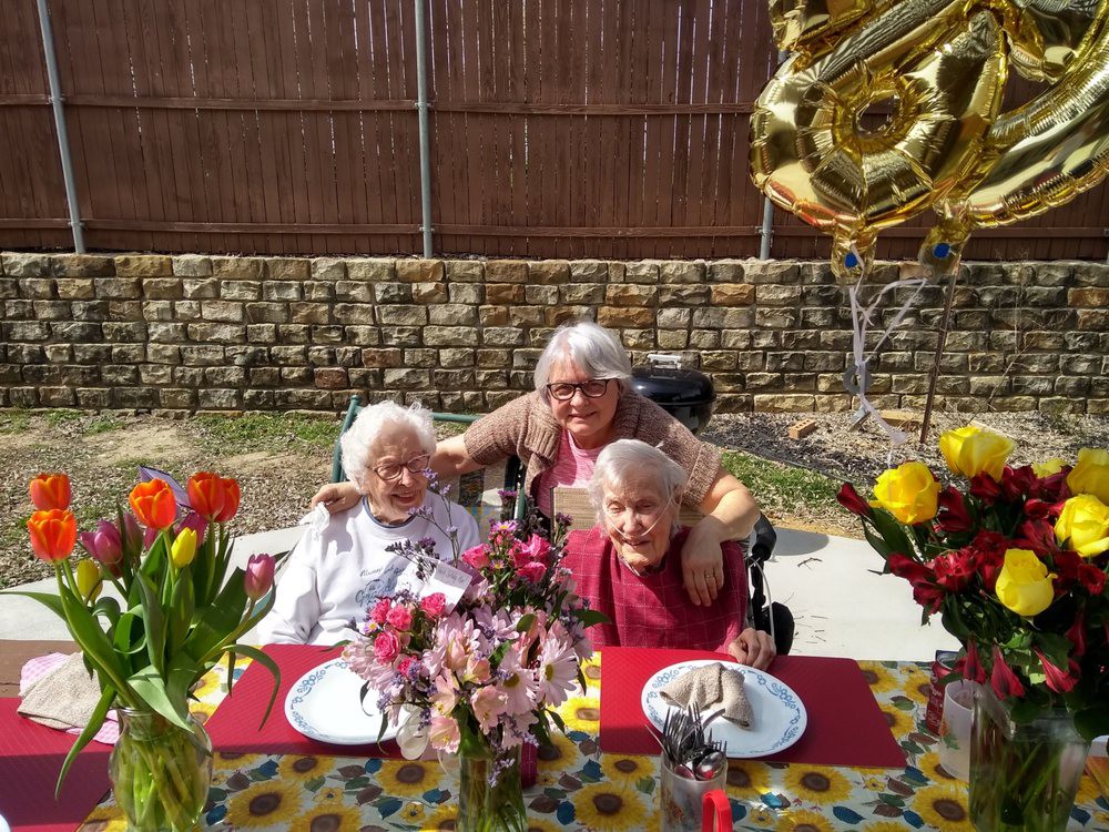 Three people celebrating outdoors with balloons and flowers at a decorated table.