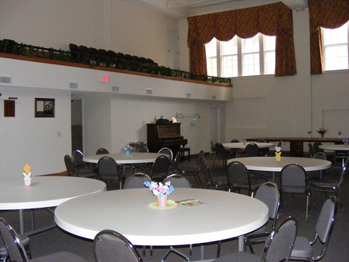 A spacious banquet hall with round tables and chairs, featuring a stage and large windows.