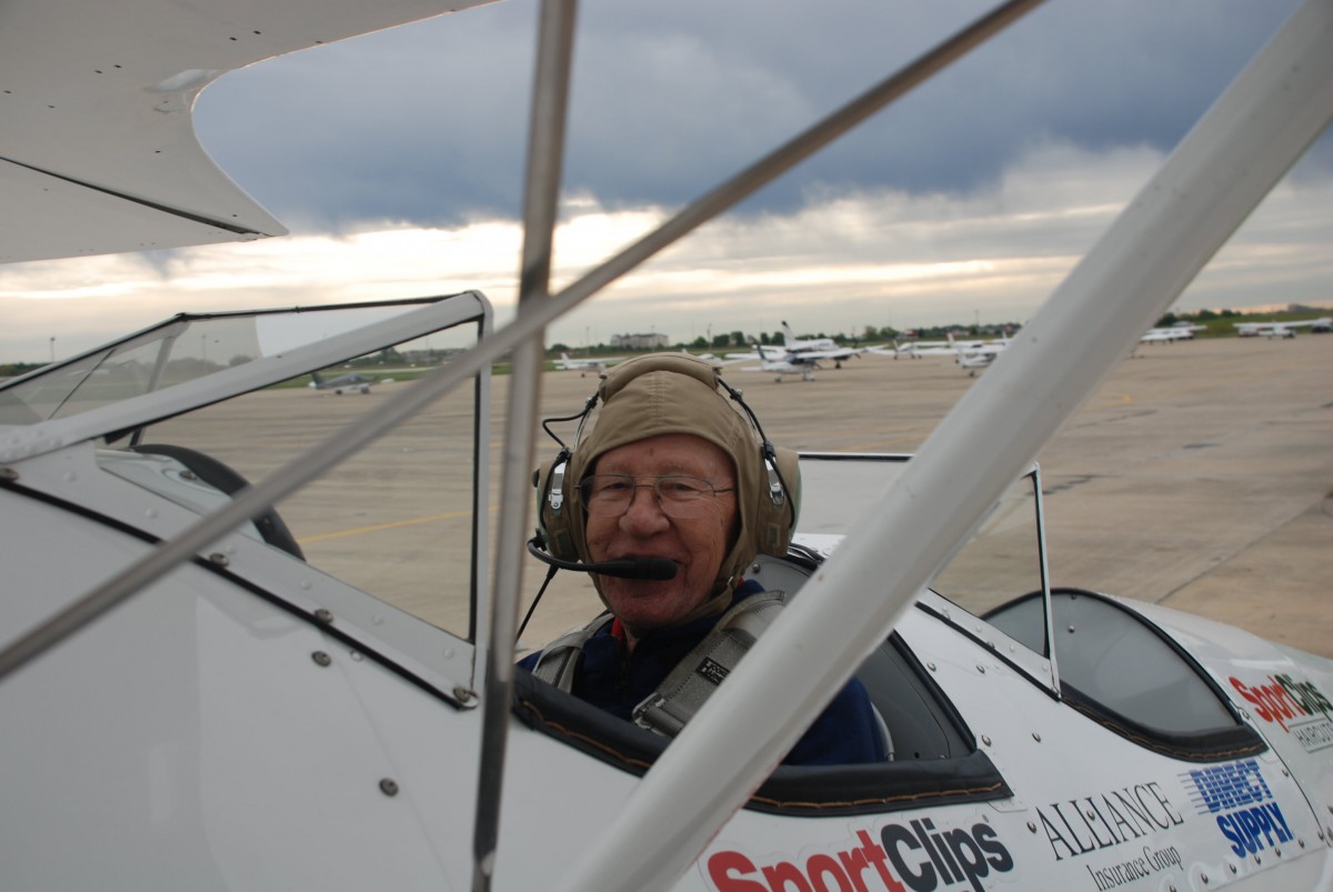 A pilot wearing a headset is seated in the cockpit of a biplane at an airfield.