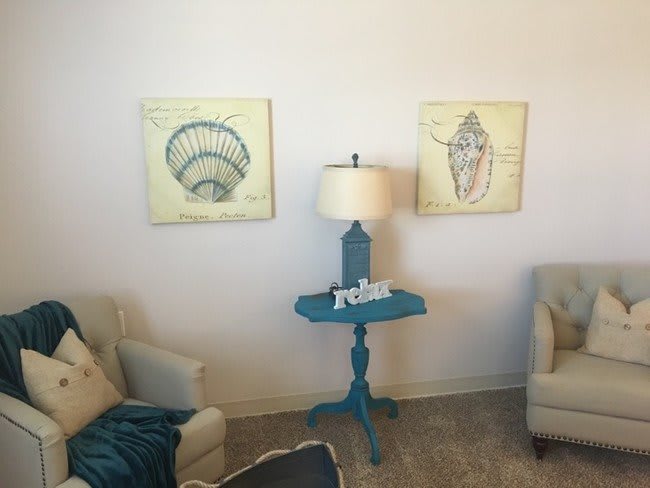 A cozy living room corner with two armchairs, a blue accent table, a lamp, and nautical artwork on the wall.