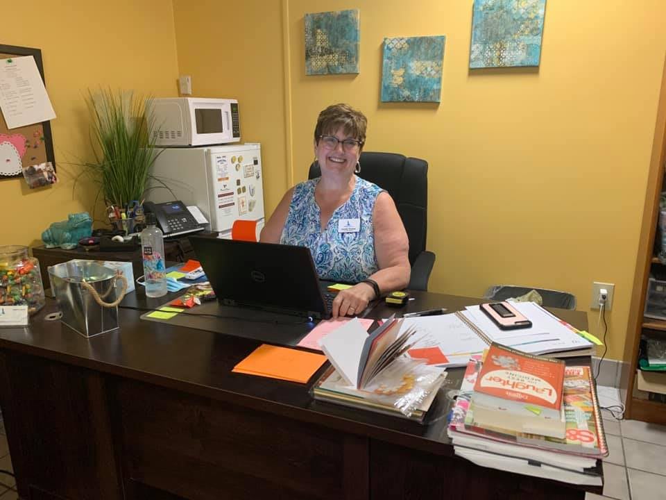 A woman sitting at a desk in an office with a laptop and various books and papers.