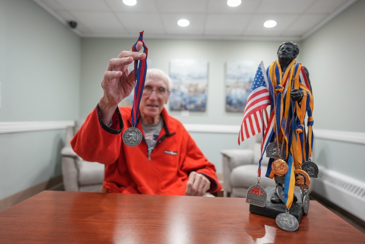 An elderly man proudly displays a medal, with a trophy adorned with multiple medals and an American flag on the table.