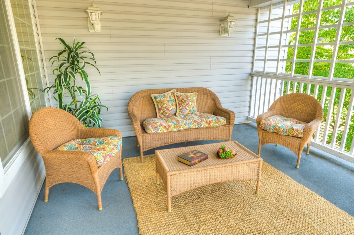 A cozy porch with wicker furniture, including a sofa and armchairs, decorated with colorful cushions and a rug.