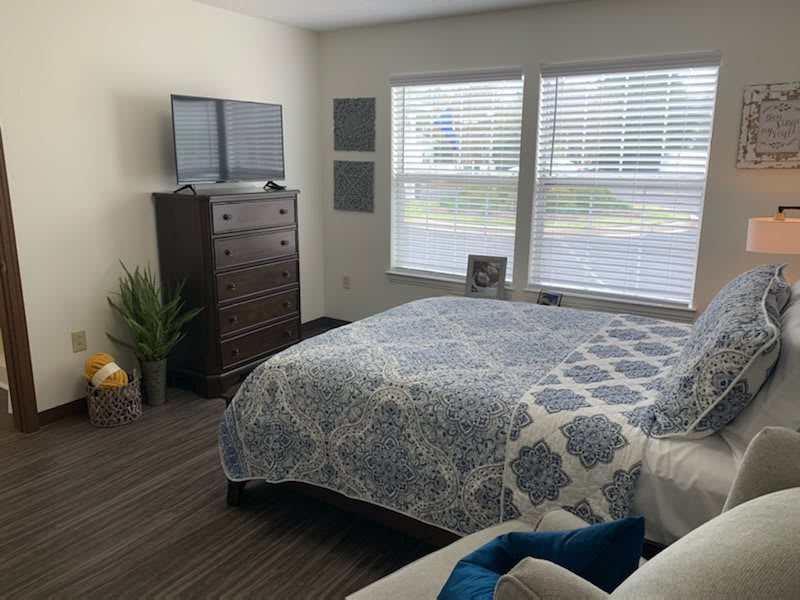 A cozy bedroom with a neatly made bed, a dresser with a television on top, and large windows providing natural light.