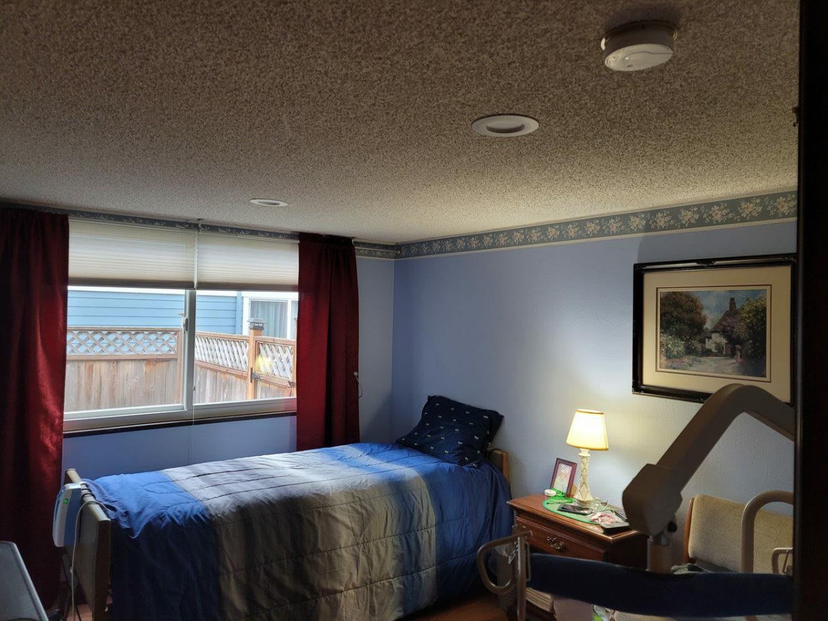 A cozy bedroom with blue walls, featuring a single bed, a window with curtains, and a nightstand with a lamp.
