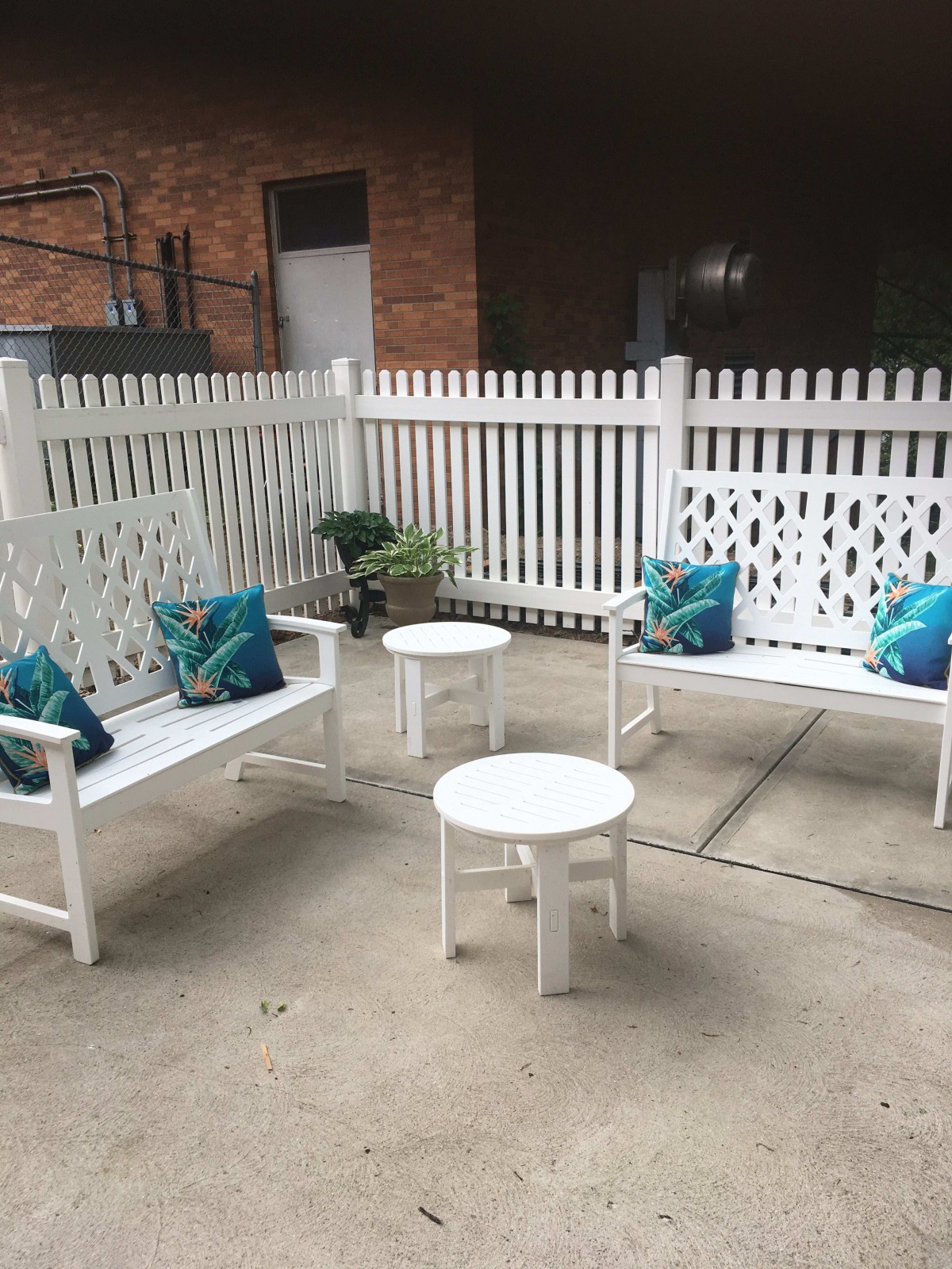A cozy outdoor patio setup with white benches, small tables, and decorative pillows in a fenced area.
