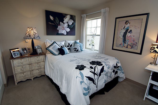 A cozy bedroom with floral-themed bedding and wall art, featuring a bedside lamp and window with curtains.