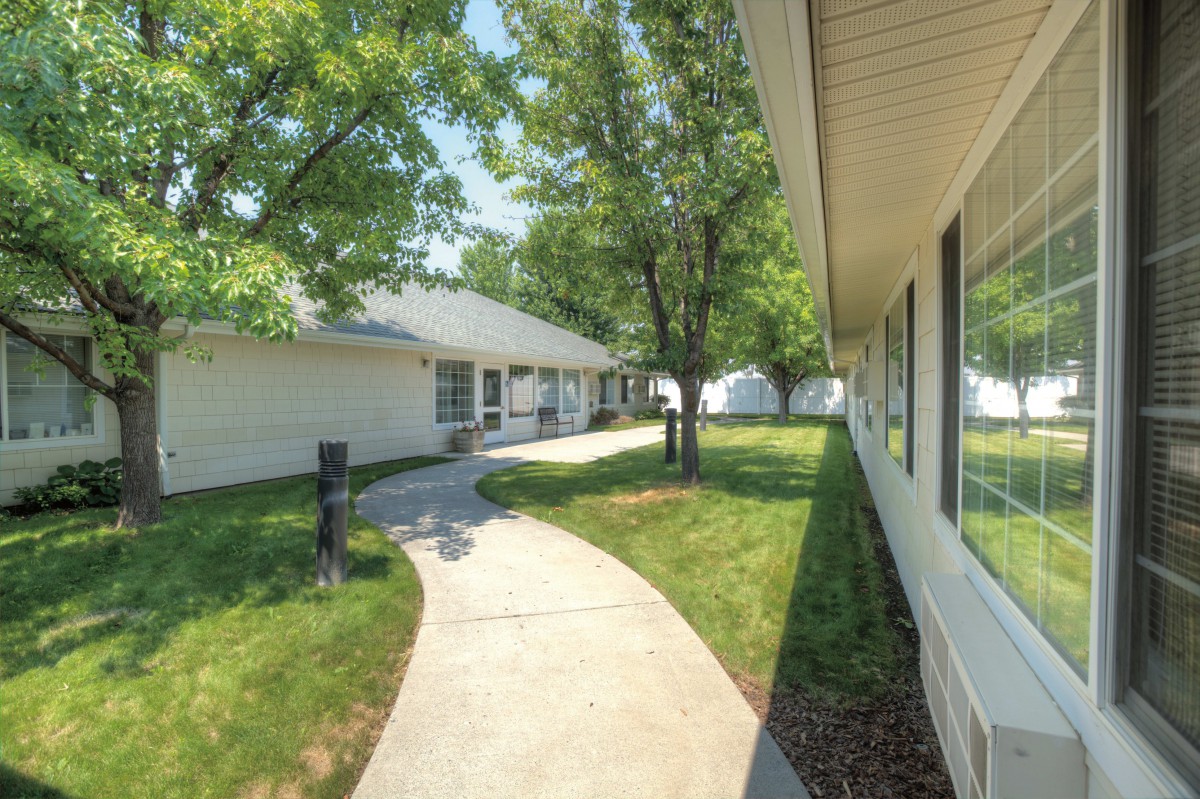 A sunny outdoor view of a residential area with white buildings, a curved sidewalk, green lawns, and trees.